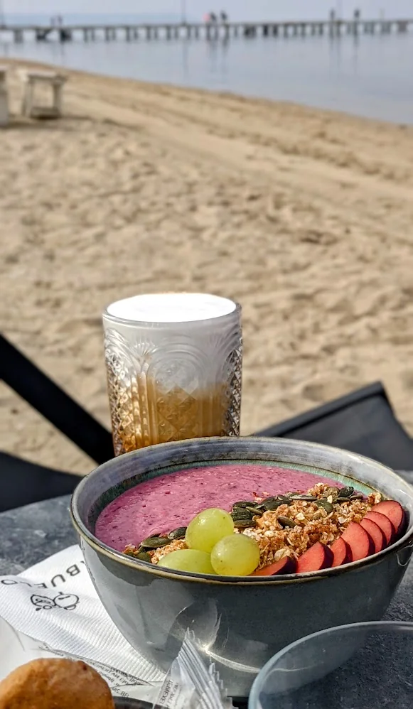 A smoothie bowl and an iced coffee on a table on the beach, with Perea’s pier behind